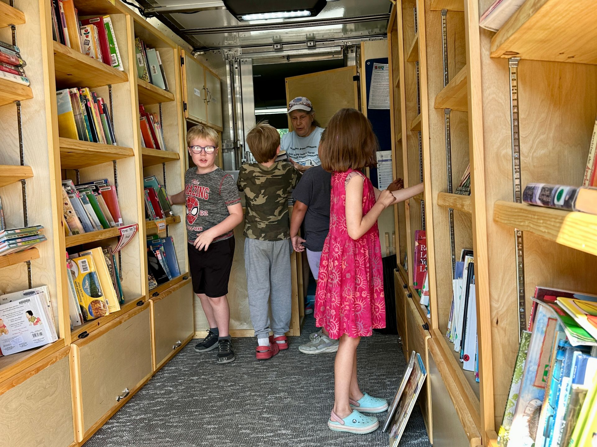 Children browsing books in a narrow library aisle, with shelves lining both sides.