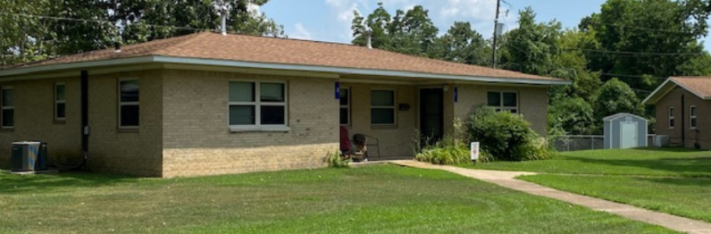 Single-story tan house with a brown roof, front windows, lawn, and a sidewalk in a suburban yard.