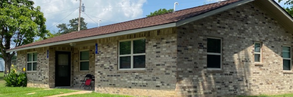 Single-story brick house with brown roof, front porch, and a few windows under a blue sky