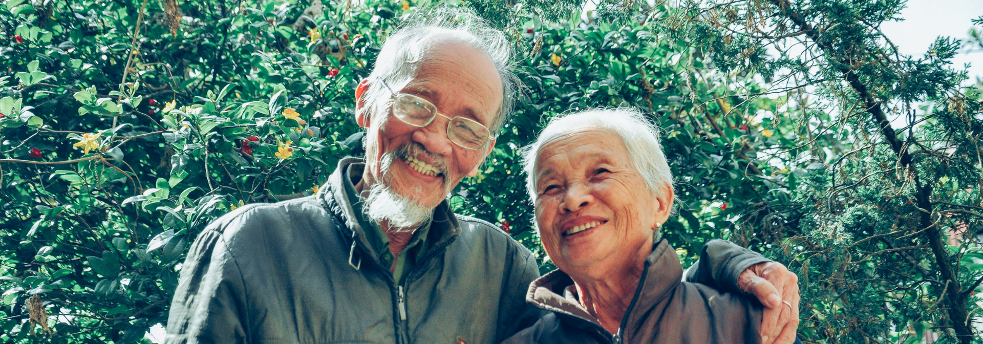 Smiling couple standing close together outdoors in front of green foliage.