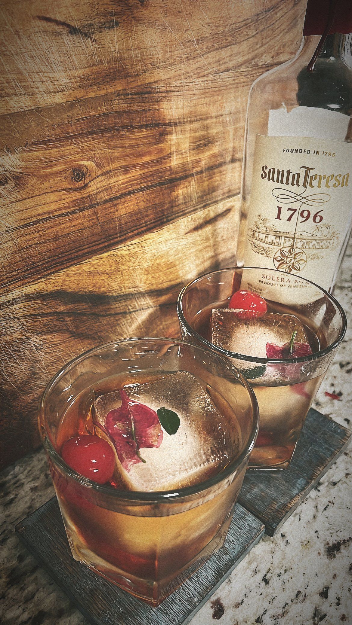 Two glasses of whiskey with cherries and ice cubes on a table.