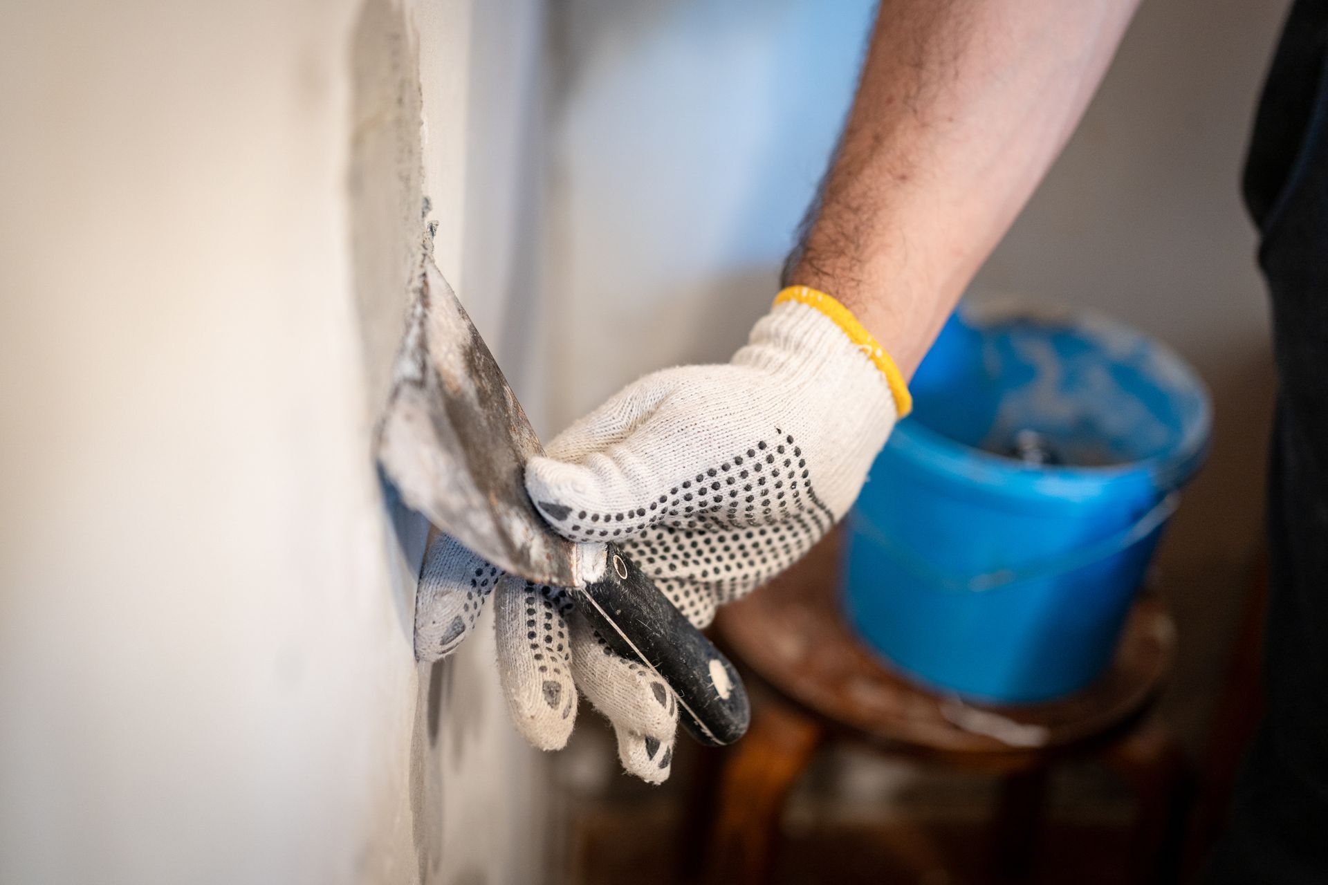 Gloved hand applying plaster to a wall with a trowel, blue bucket in the background