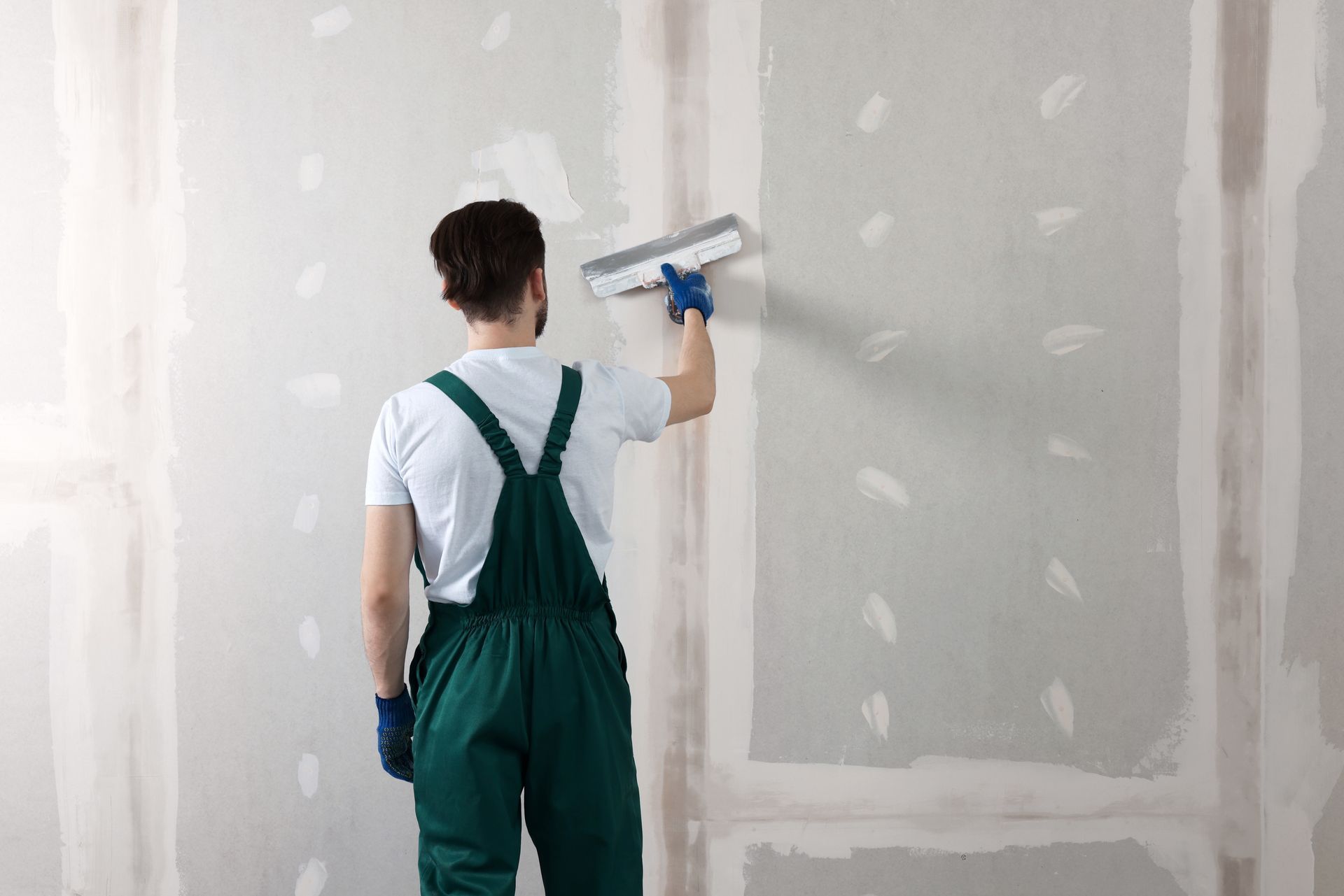 Person in green overalls and blue gloves smoothing white drywall with a trowel in a room under renovation