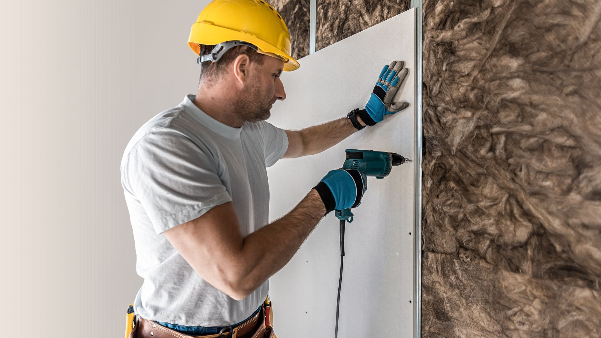 Construction worker installing drywall with a power drill, wearing a yellow hard hat and gloves.