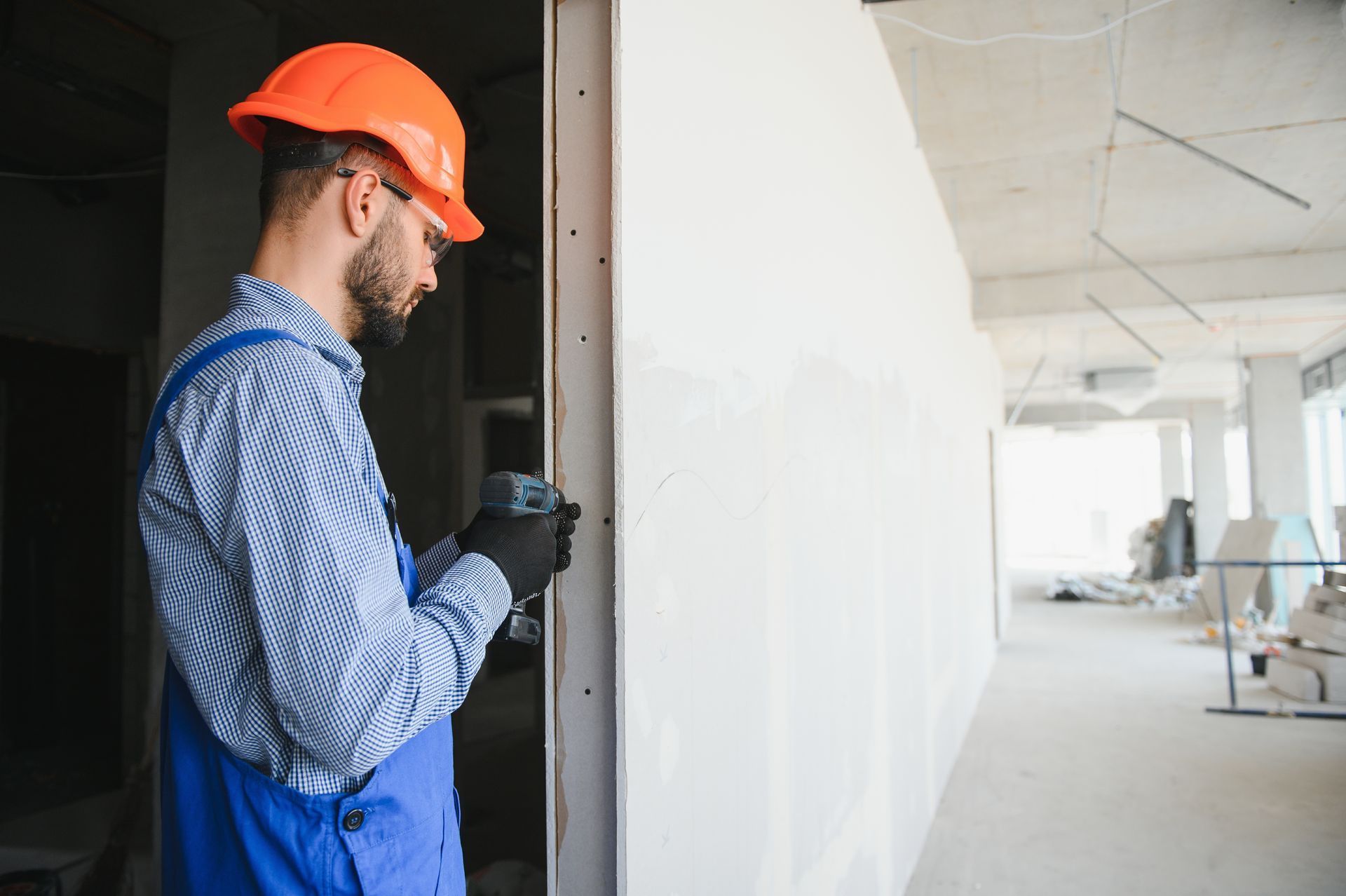 Construction worker in orange hard hat and blue overalls standing by a doorway in a white hallway