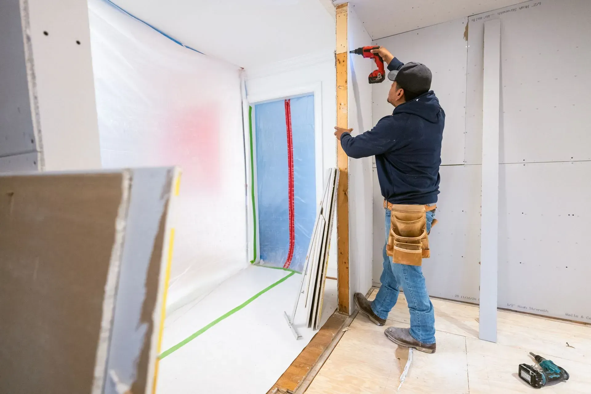 Worker installing drywall in a room, using a drill near a doorway with protective coverings on the floor.