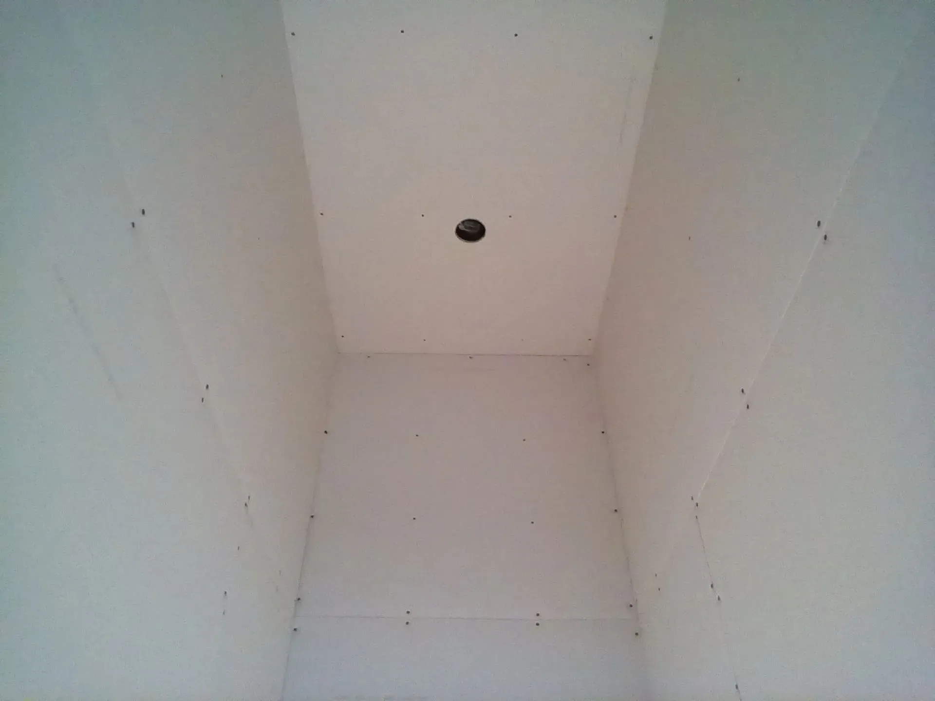 White drywall interior of a narrow rectangular shaft or closet, with a small black hole in the ceiling.