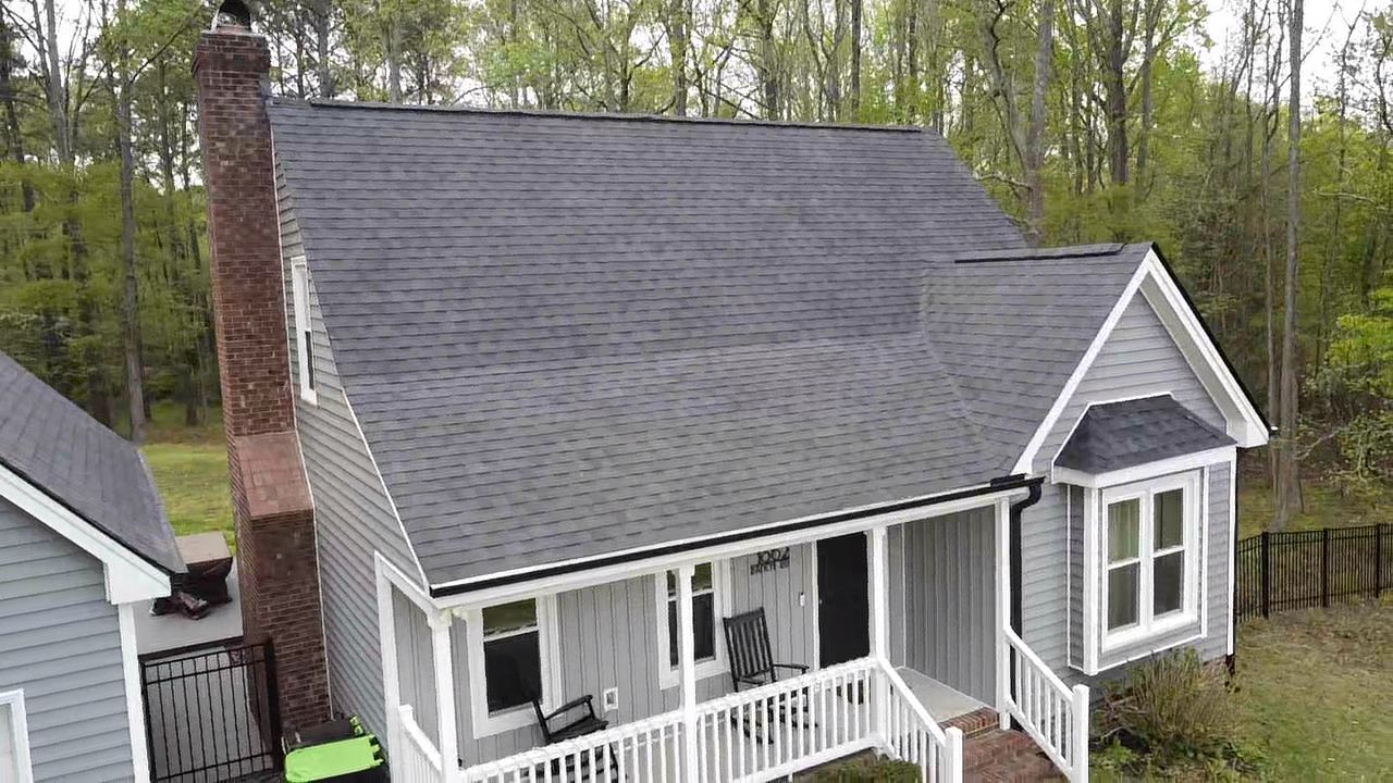 Gray house with porch, chimney, and dark roof, surrounded by trees.