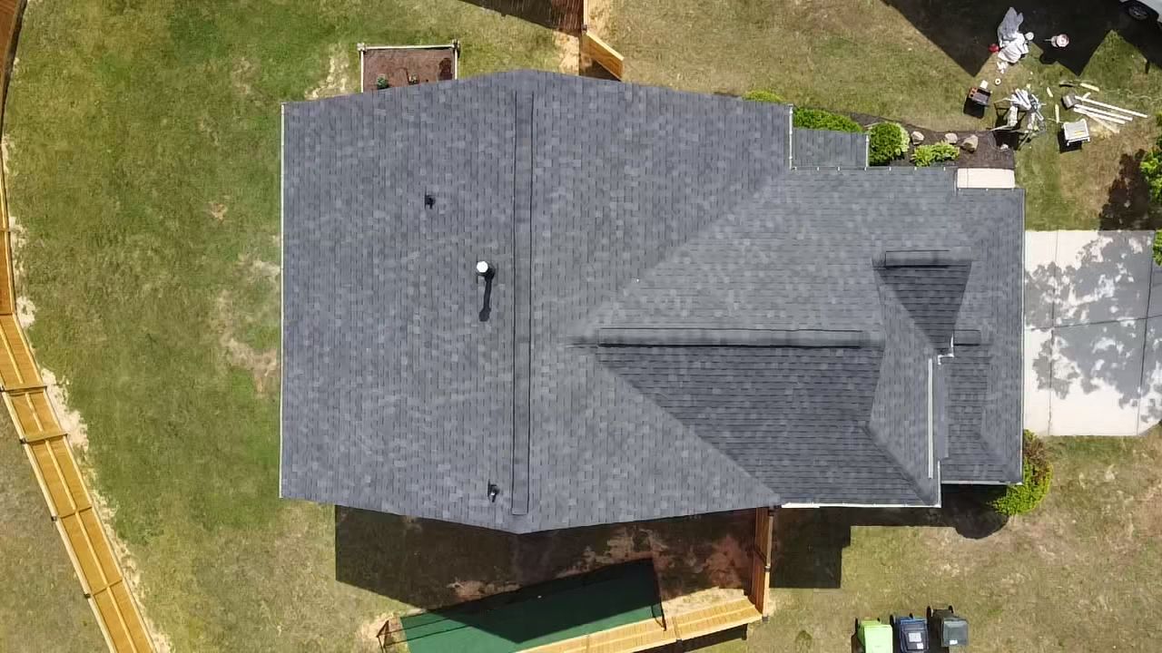 Overhead view of a house with a dark gray roof on green grass; wooden fence, bushes, and driveway visible.