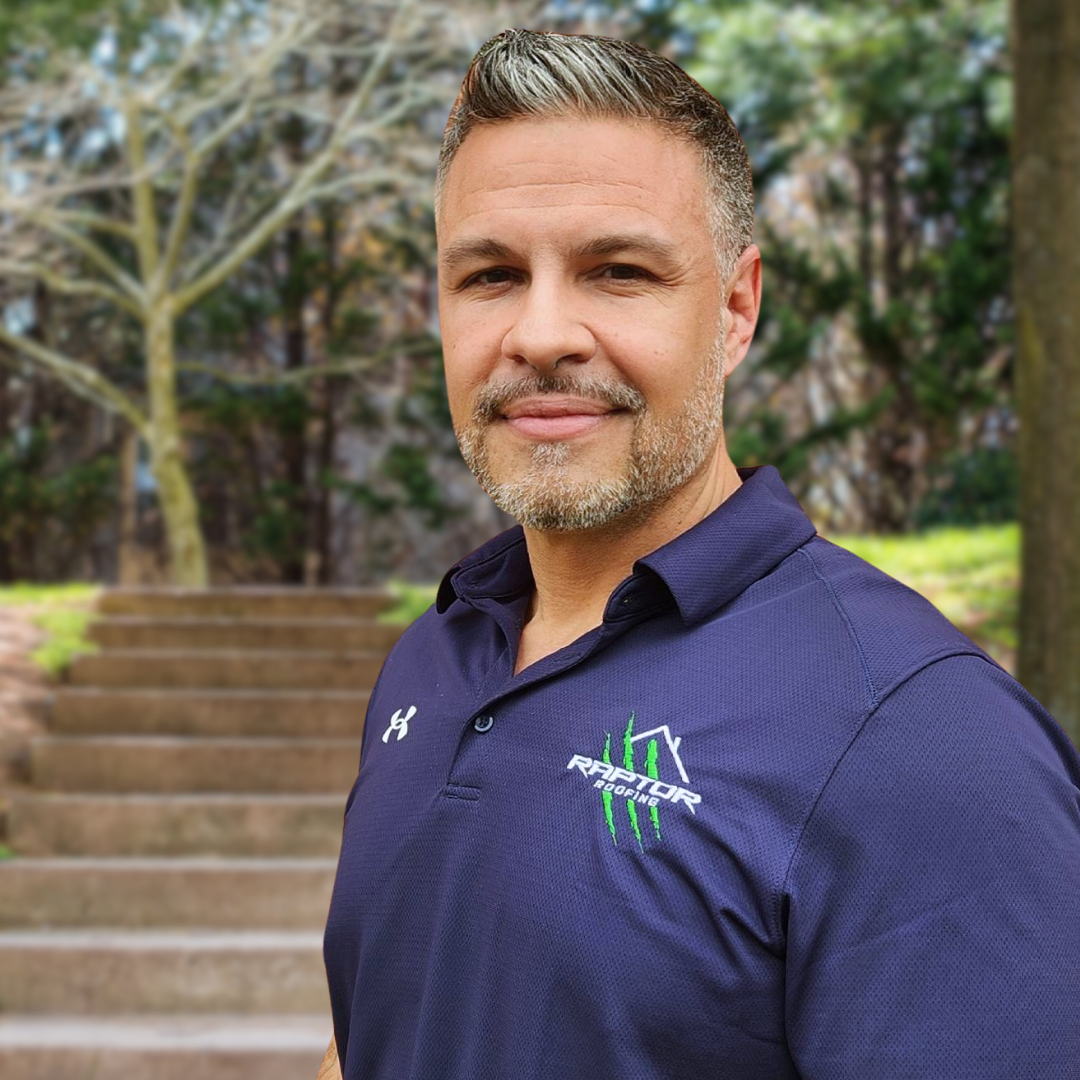 Man with graying hair and beard smiles, wearing navy polo shirt with Raptor logo, outdoors by stone steps.