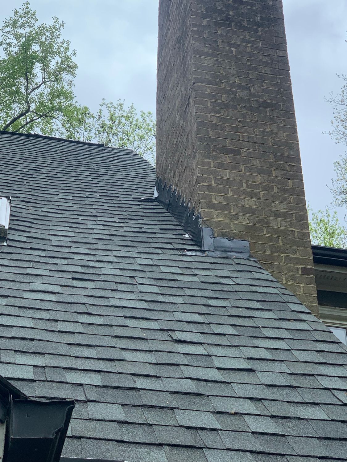 Gray shingle roof with a brick chimney. Green trees in the background under a cloudy sky.