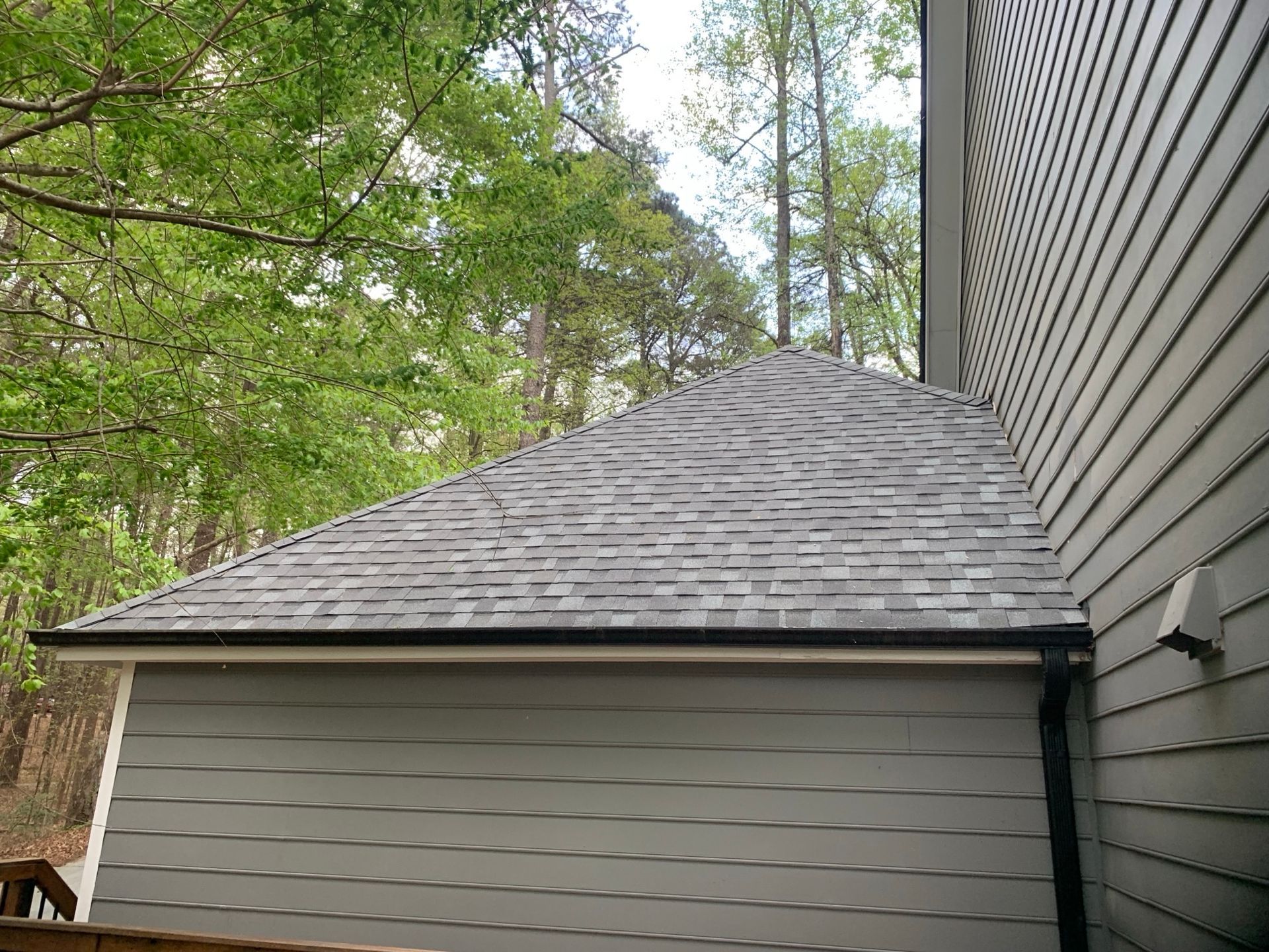 Gray shingled roof with a gutter, attached to a gray-sided building, with trees in the background.