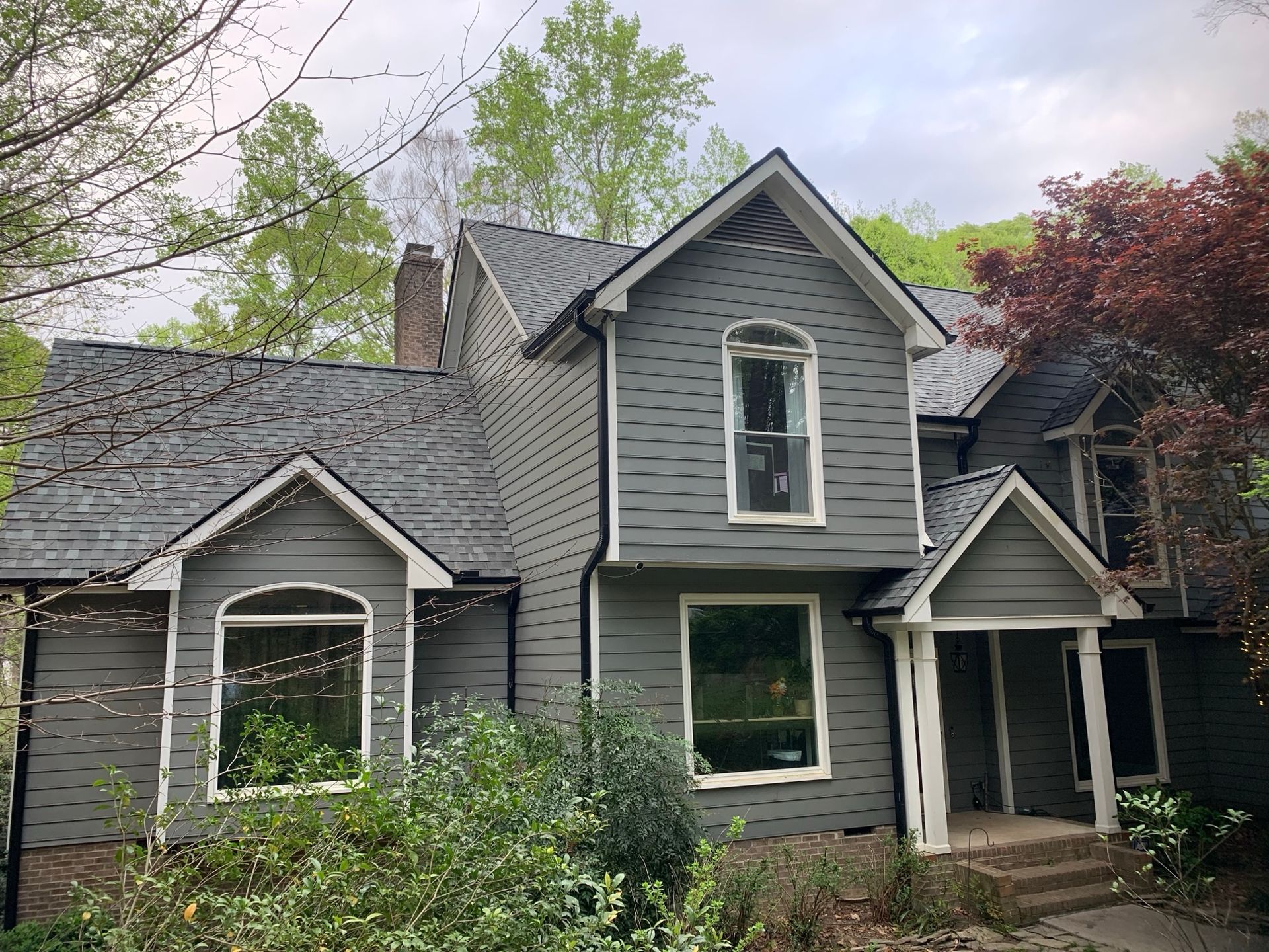 Gray two-story house with a gray roof and white trim, surrounded by green trees and bushes.