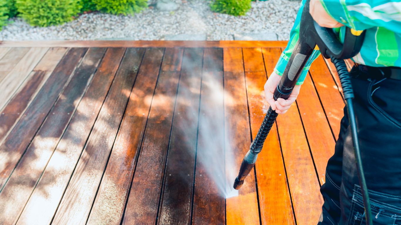 Person pressure-washing a wooden deck, spraying water across orange-brown boards