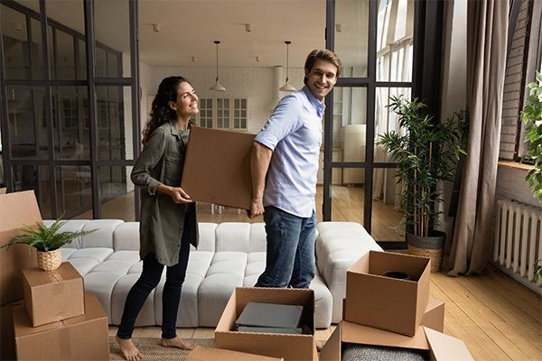 Picture of two people carrying a moving box with more moving boxes around them in the living room