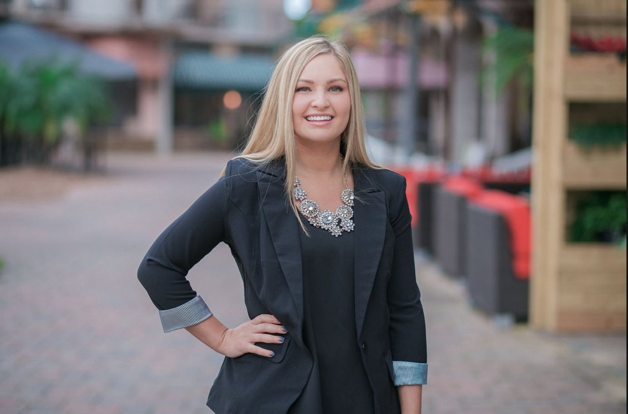 Woman in black blazer smiles, poses outdoors with hand on hip.