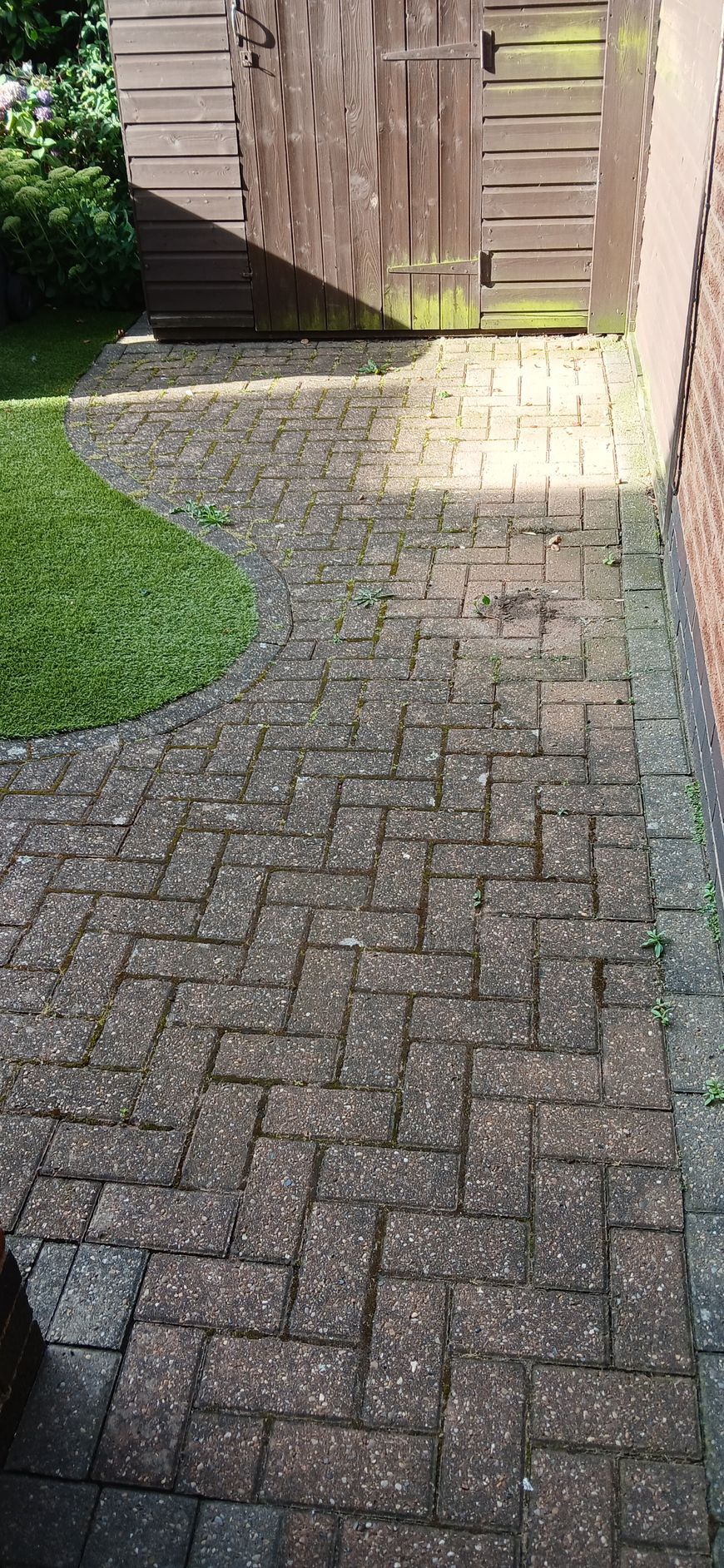 A block paved patio leading to a shed next to a lawn area before being pressure washed.