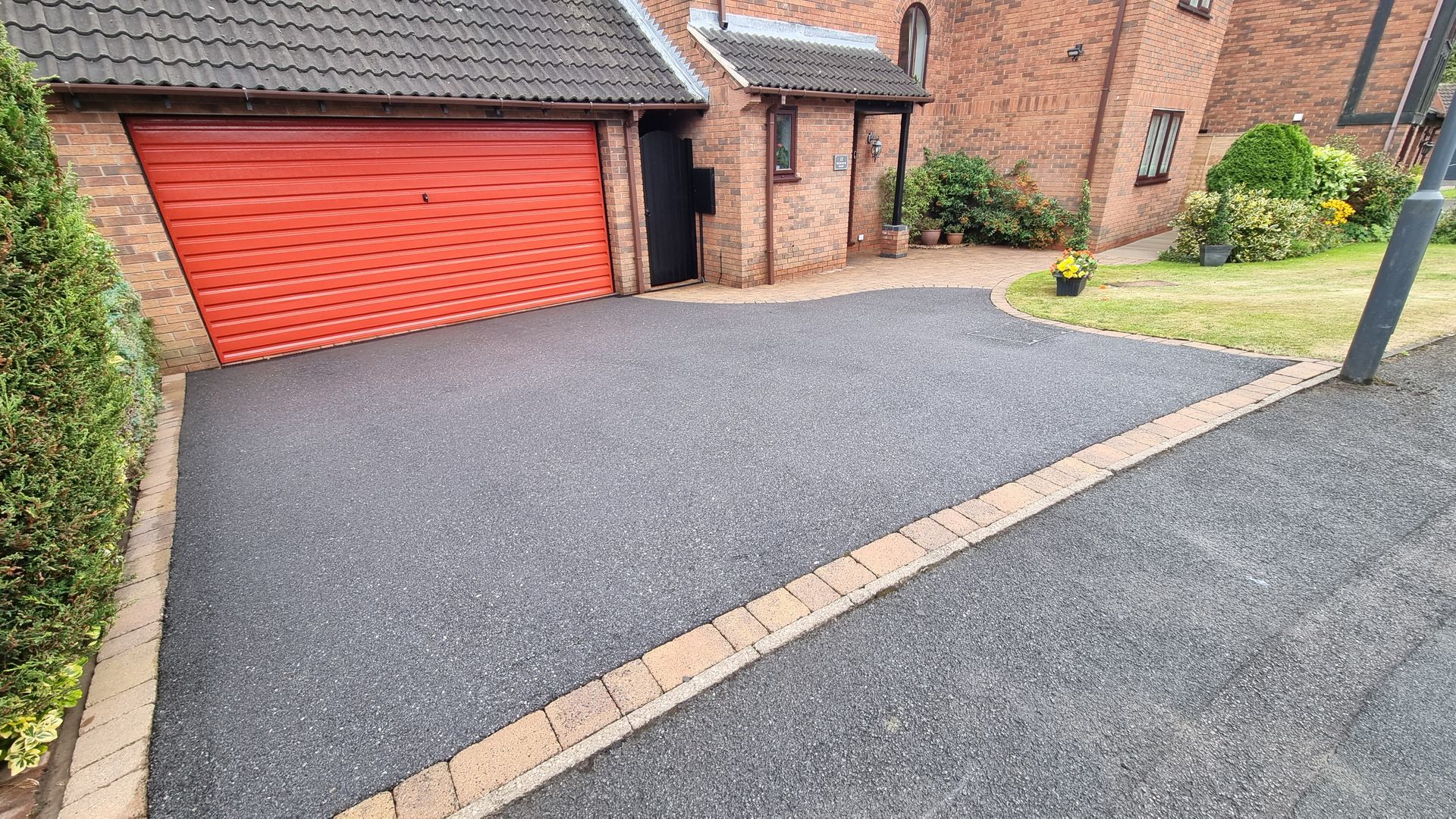A tarmac driveway with a red garage door in front of a brick house, before being cleaned.