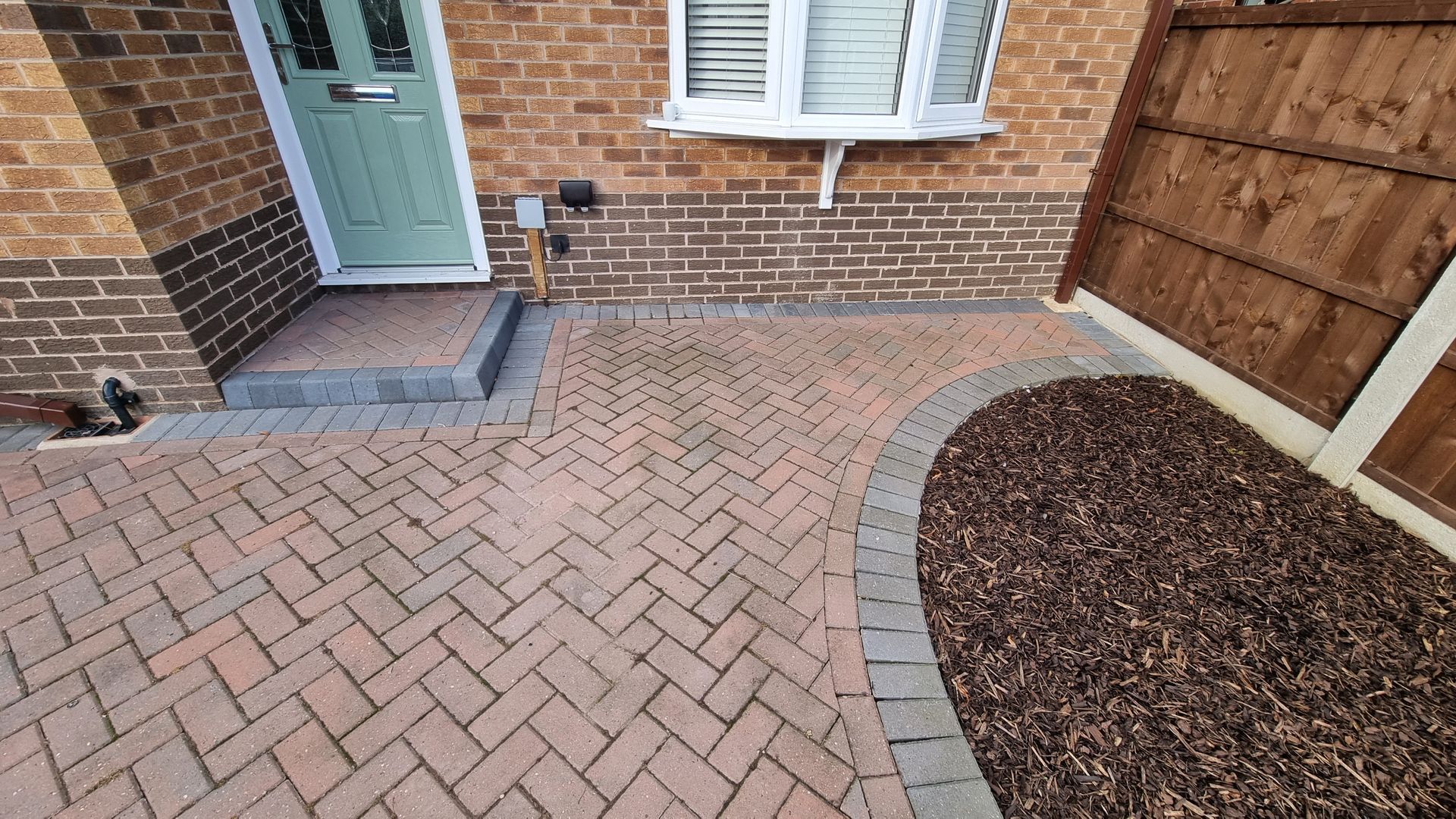 A brick driveway in front of a brick house with a wooden fence.