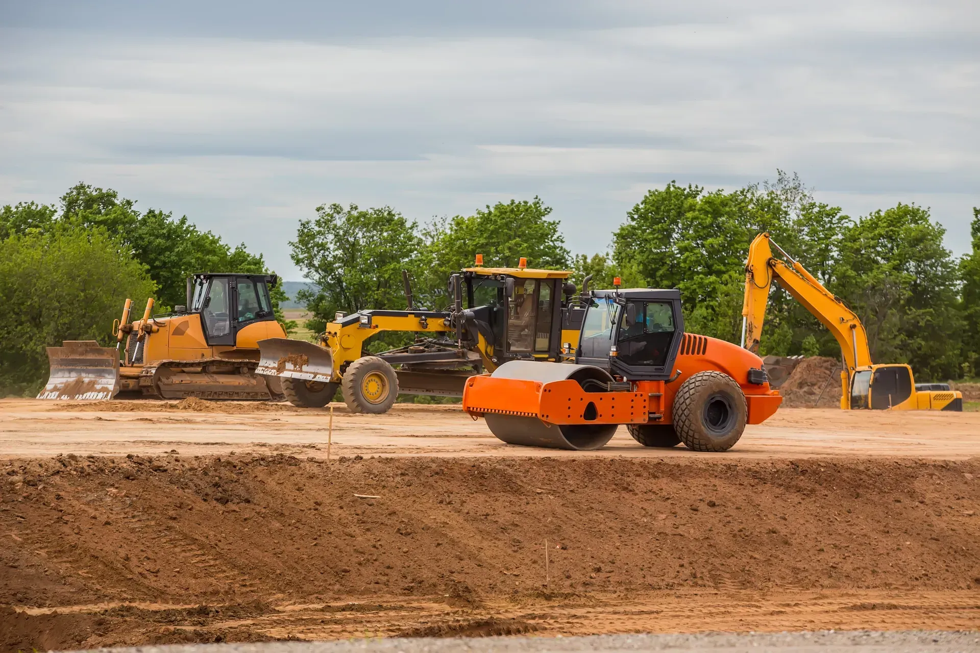 Construction vehicles on a dirt site, including bulldozers, a loader, and an excavator under cloudy skies