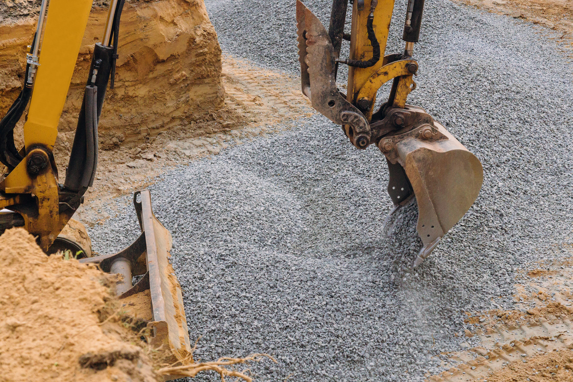 Excavator bucket spreading gravel beside a mound of sand at a construction site