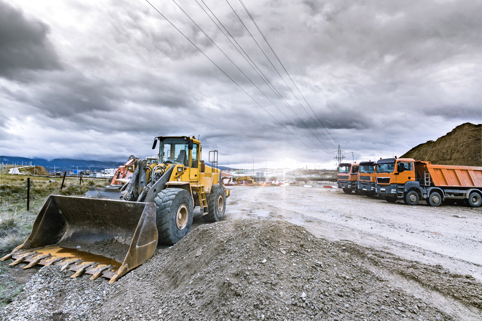Yellow bulldozer and orange dump trucks on a muddy construction site under cloudy skies