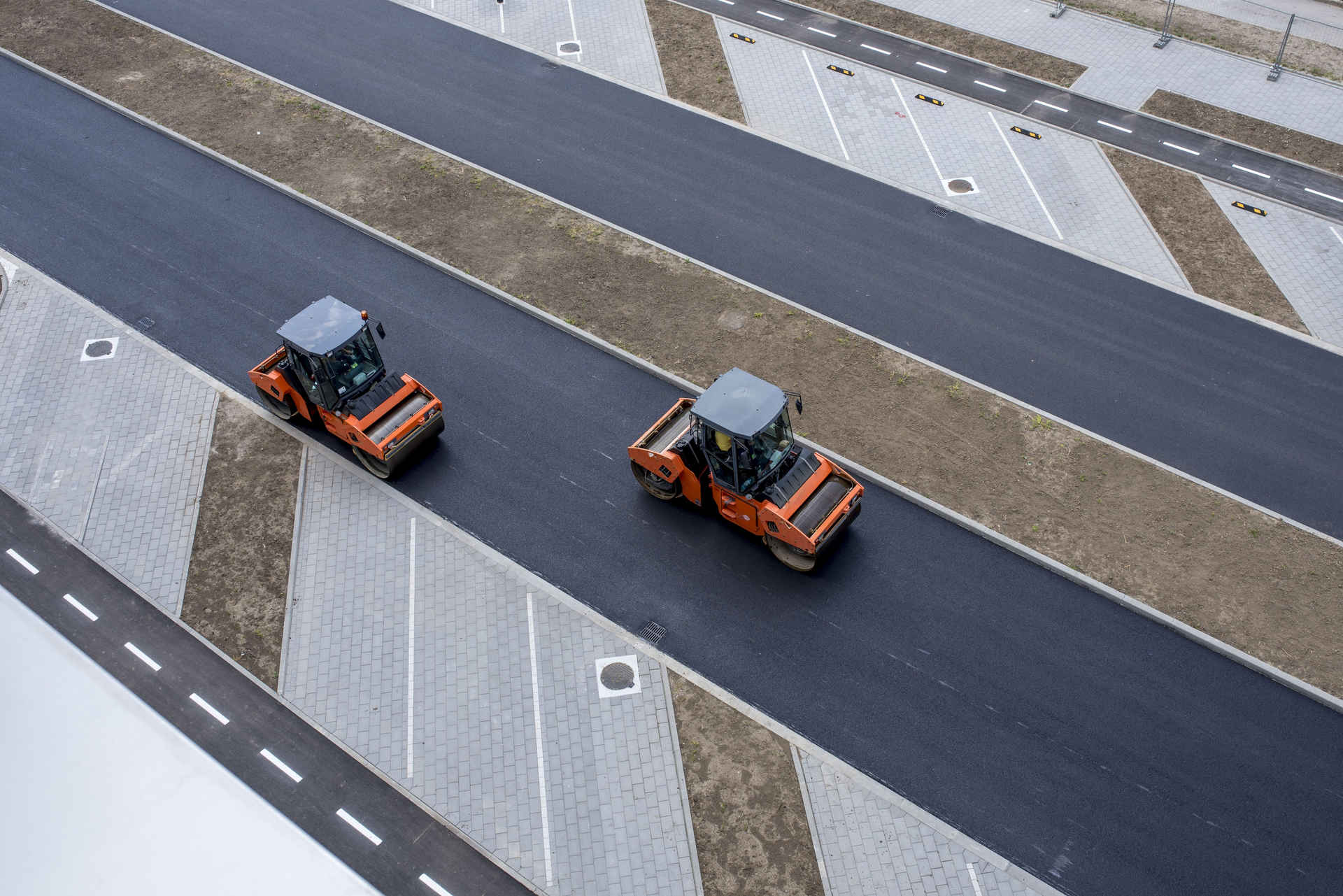 Two orange tractors driving on a wide paved road beside gray striped pavement