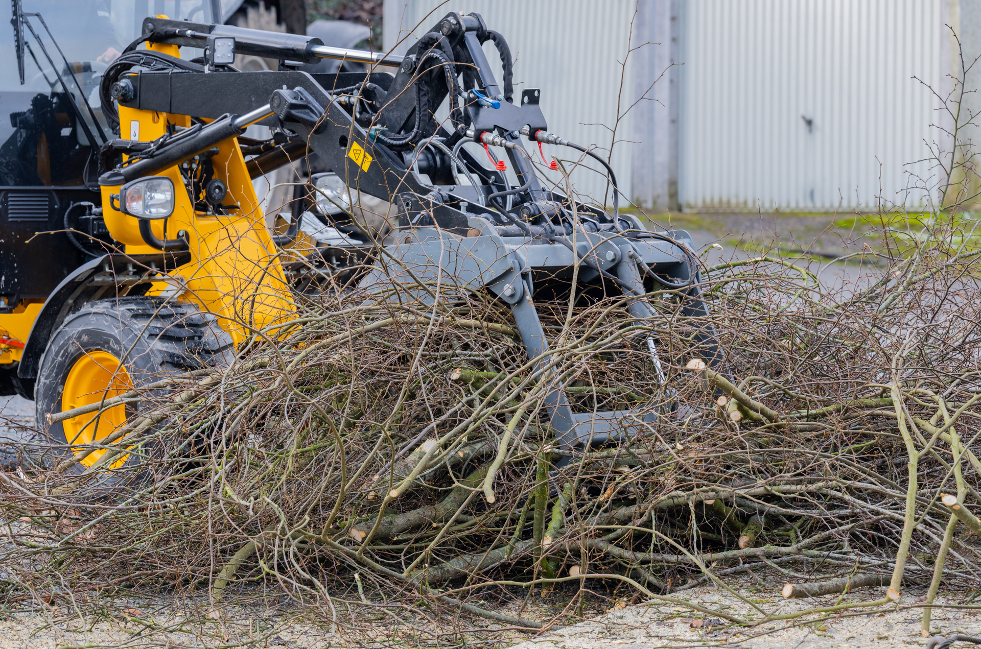 Yellow skid-steer loader clearing brush and branches beside a building