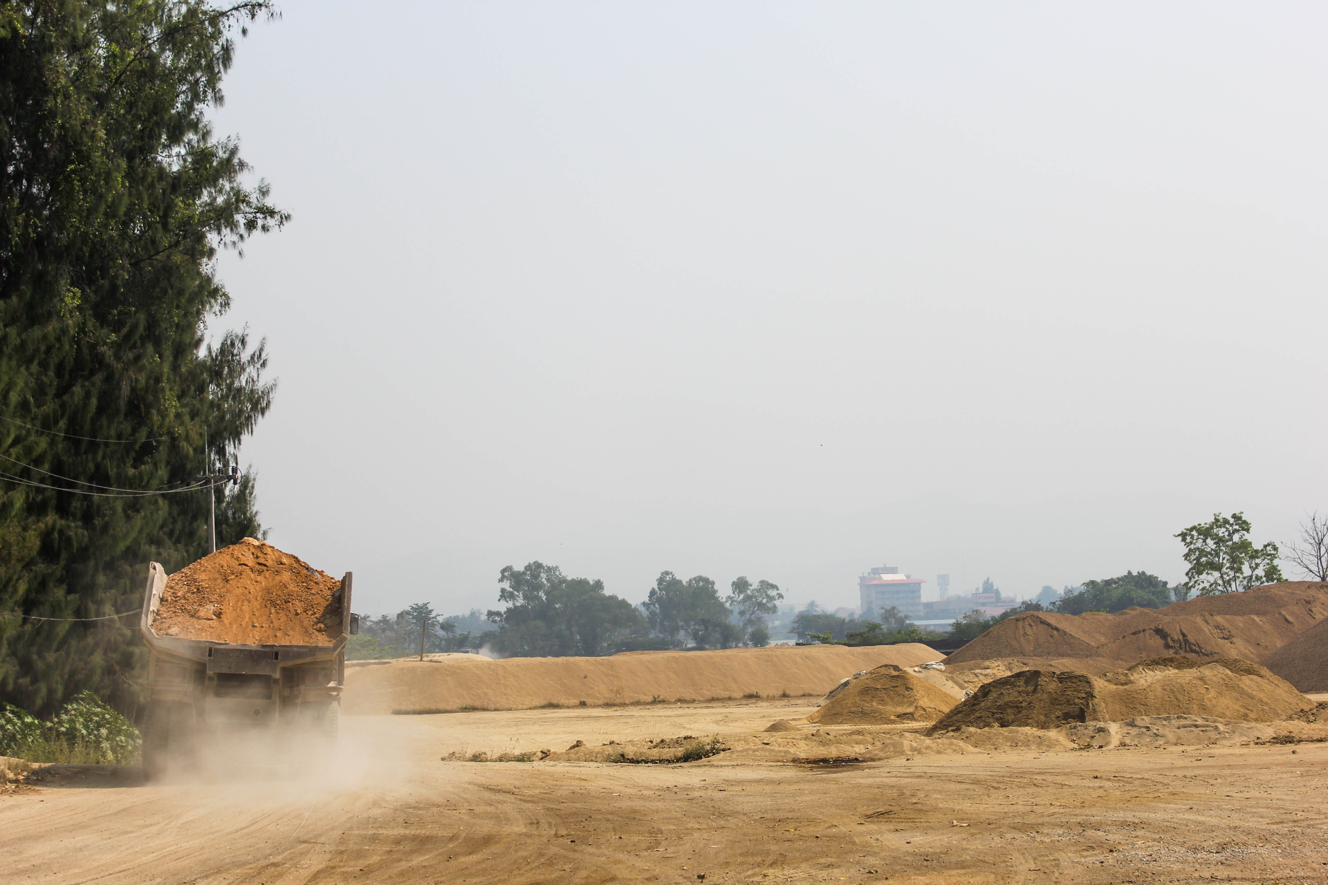 Dusty construction site with a dump truck unloading soil beside piles of dirt and trees