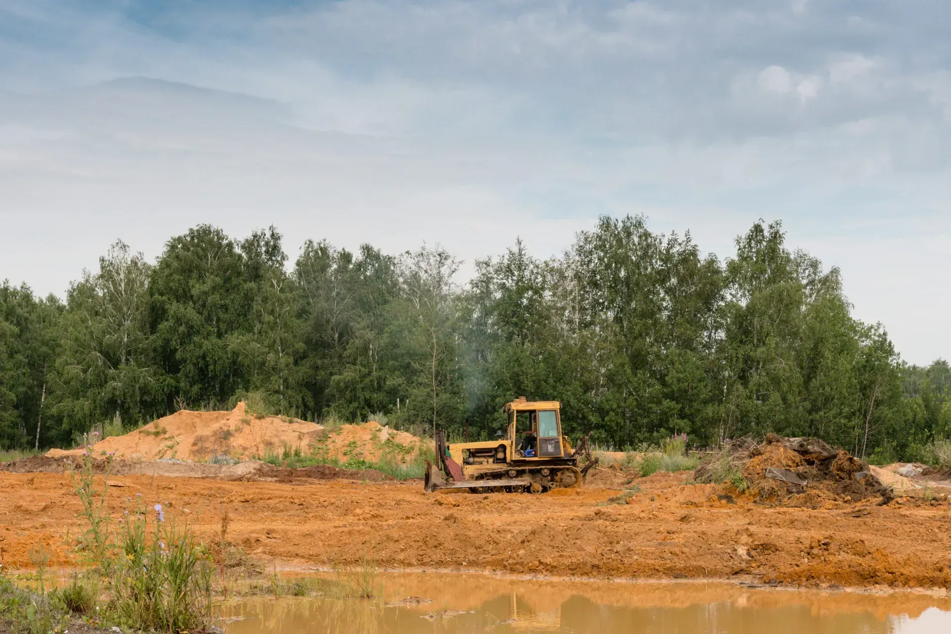 Bulldozer clearing red dirt on a muddy construction site beside a tree line under a cloudy sky
