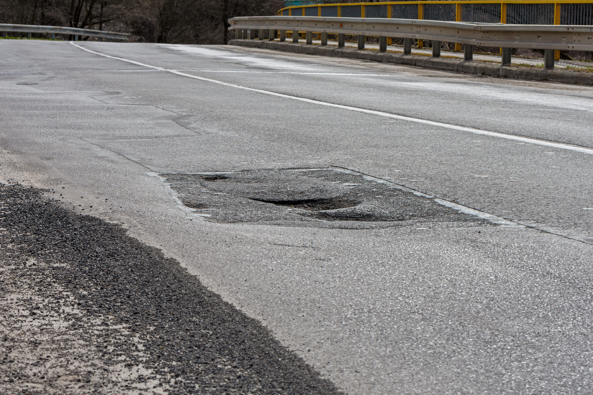 Damaged road with a large pothole and rough asphalt near a guardrail.
