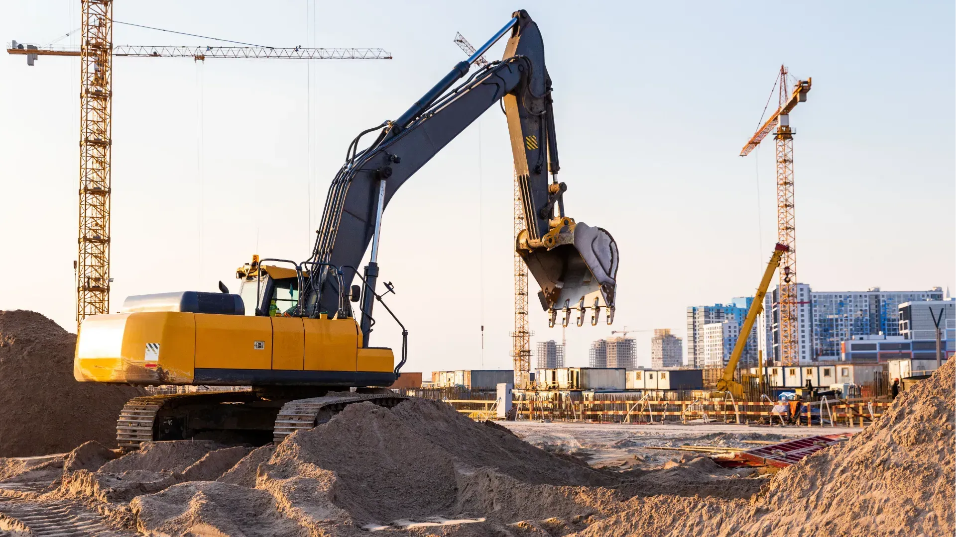 Yellow excavator lifting soil at a construction site with cranes in the background