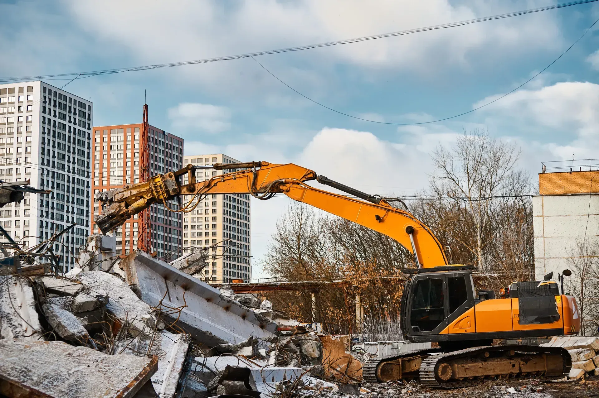 Orange excavator demolishing concrete rubble at a construction site with apartment buildings in the background
