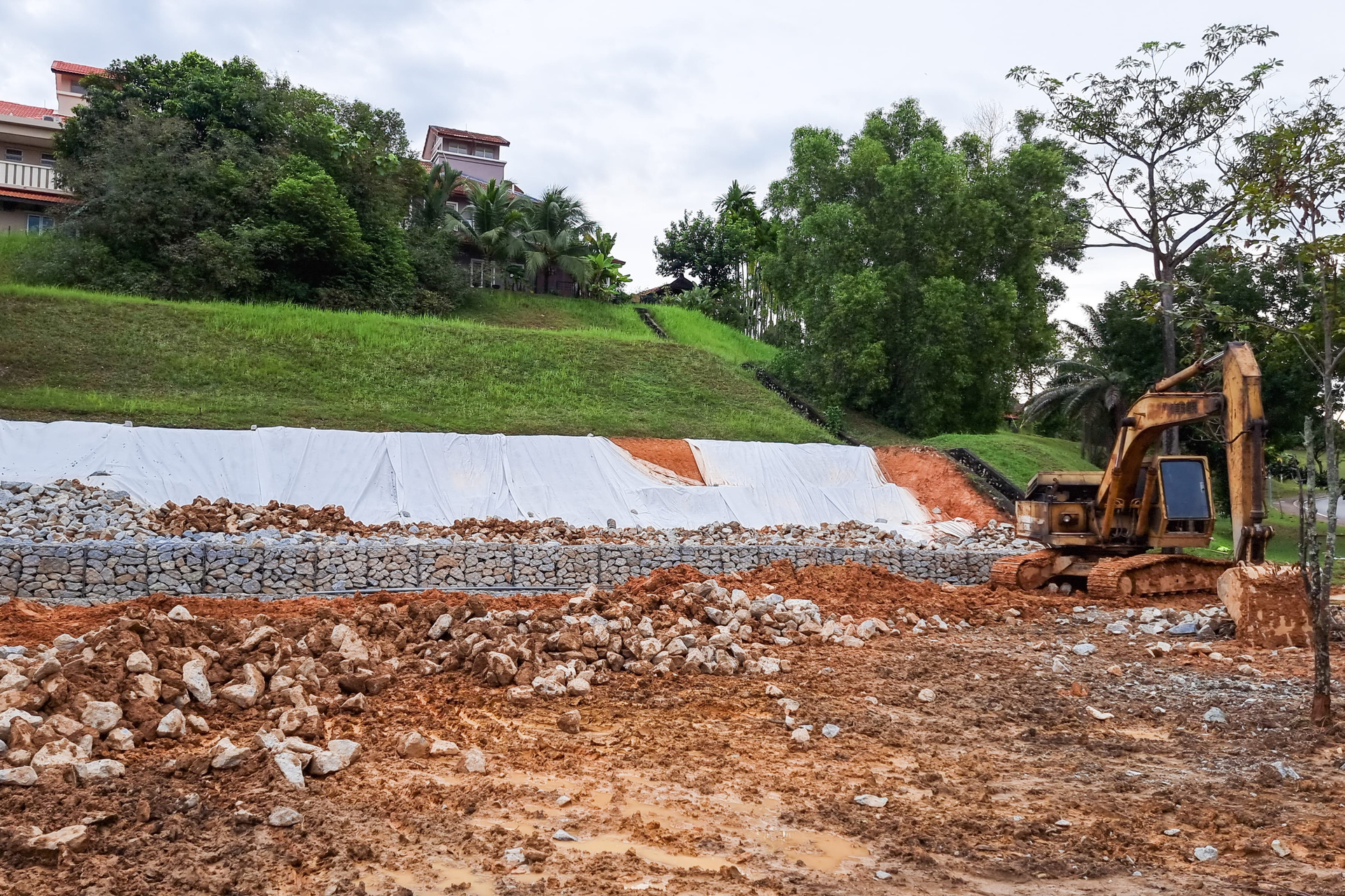 Bulldozer clearing muddy construction site beside a sloped hillside and green trees.