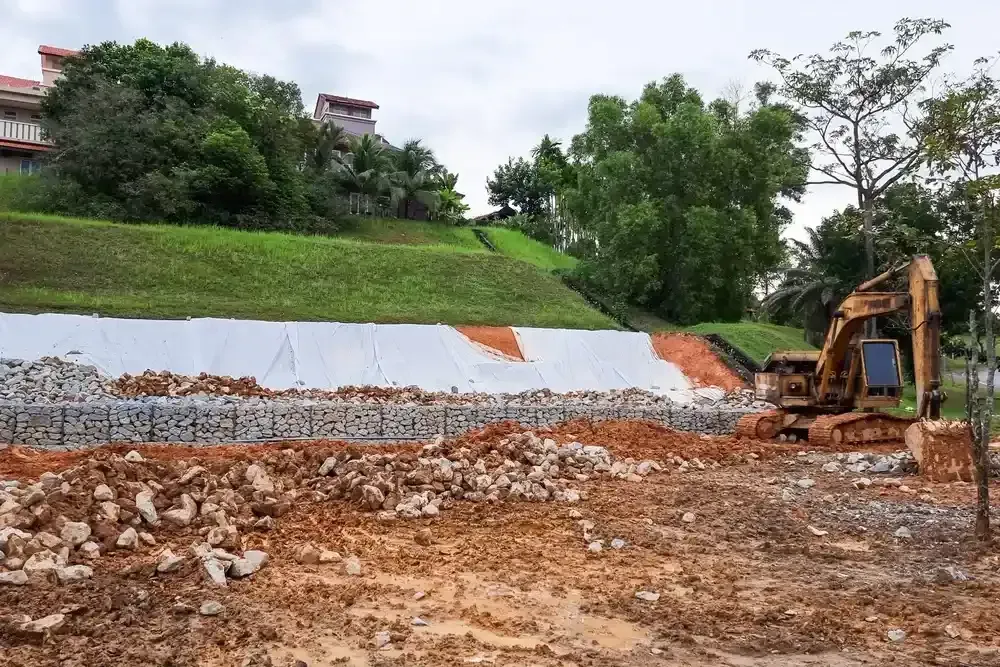 Excavator working on a muddy construction site beside a sloped retaining wall and green hillside.