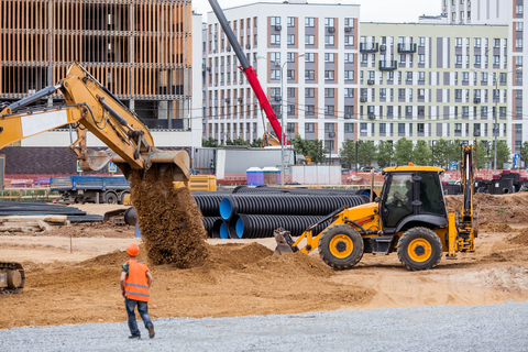 Construction site with excavator dumping soil beside a loader and worker in orange vest