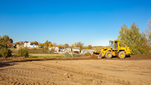 Yellow bulldozer grading a dirt lot under a clear blue sky, with houses and trees in the background