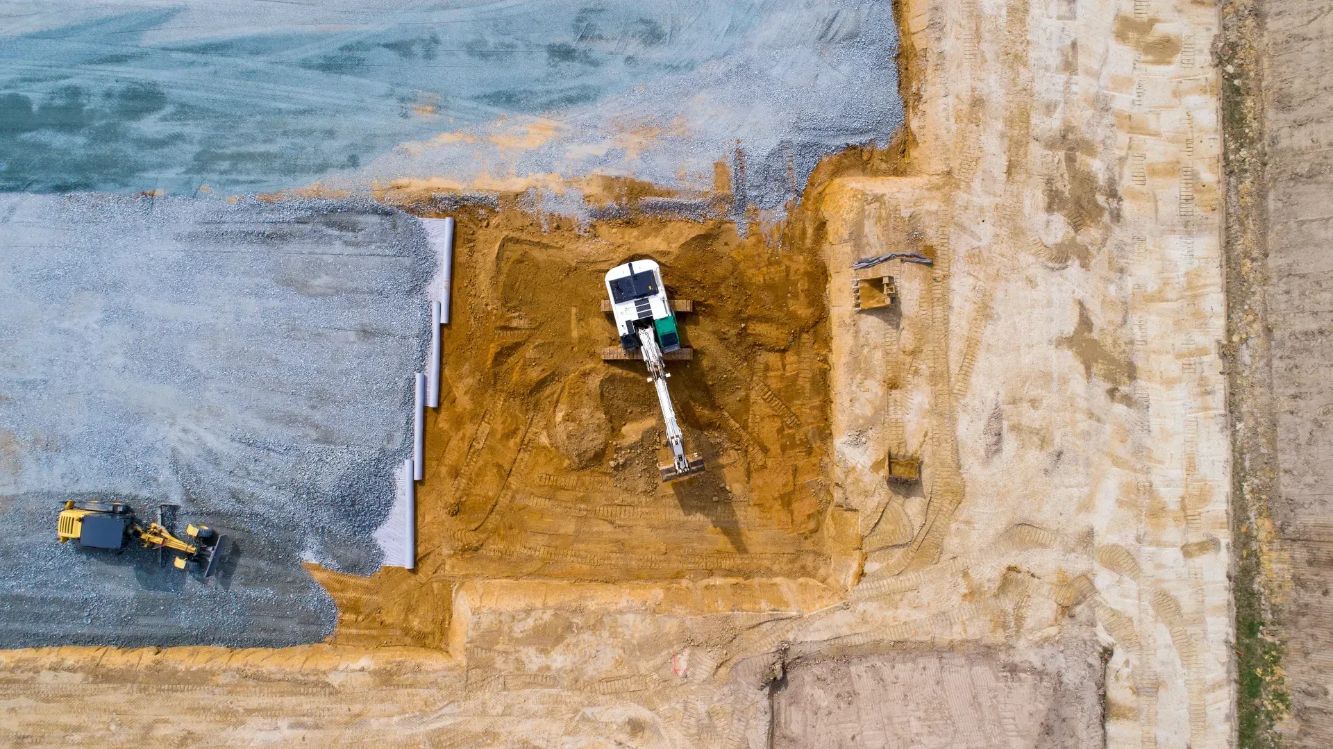 Aerial view of two construction vehicles grading a dirt lot beside a gray paved area.