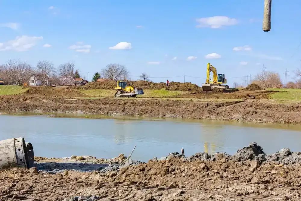 Two excavators working on a muddy riverbank beside calm water under a blue sky