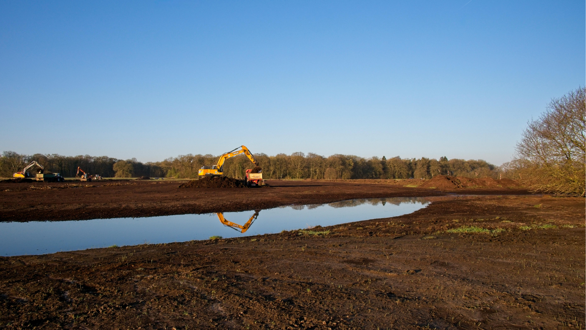 Excavator working beside a muddy pond in a rural dirt lot under a clear blue sky