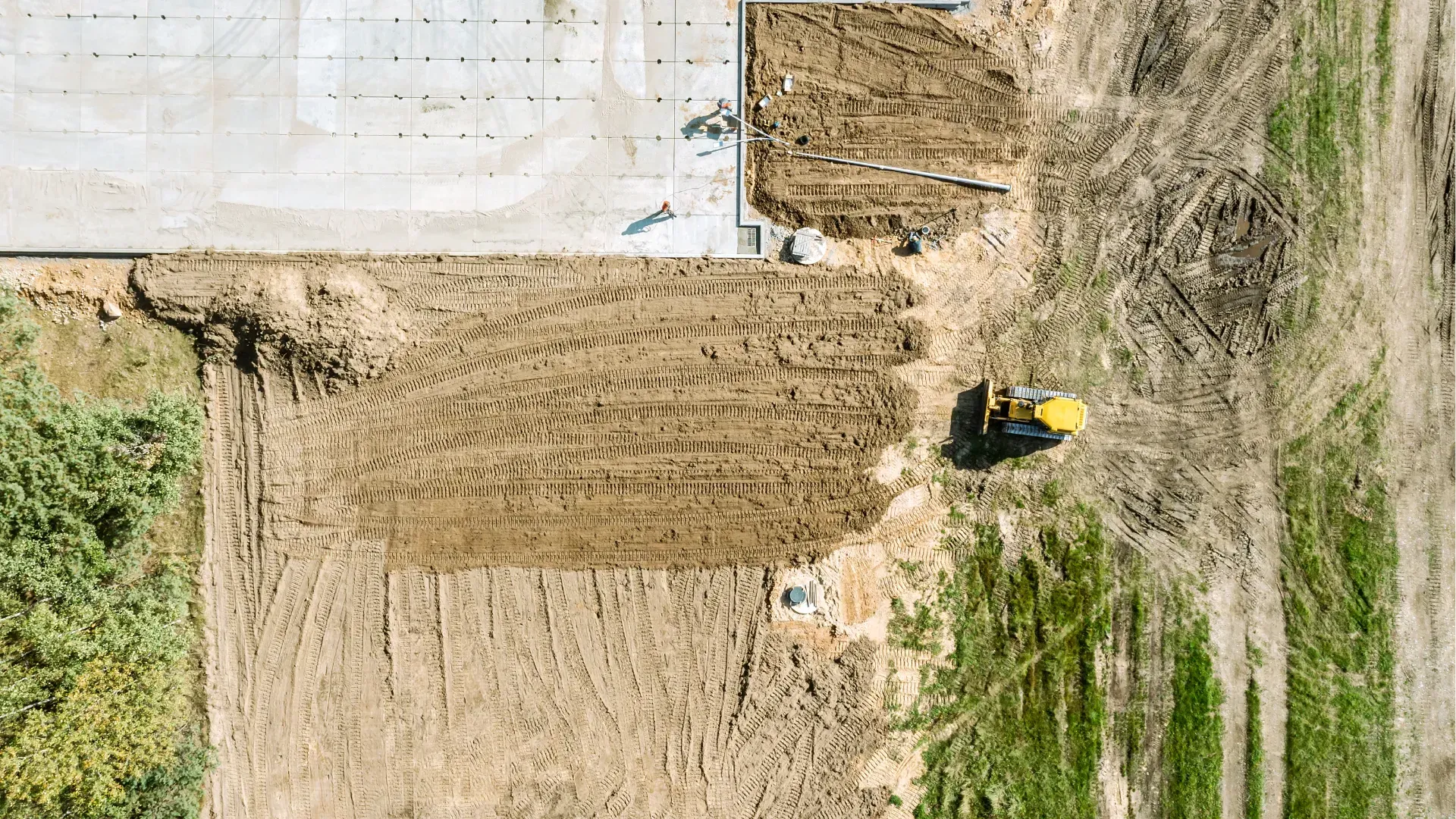 Person in a yellow helmet operating a tractor on a dirt lot beside a white building.