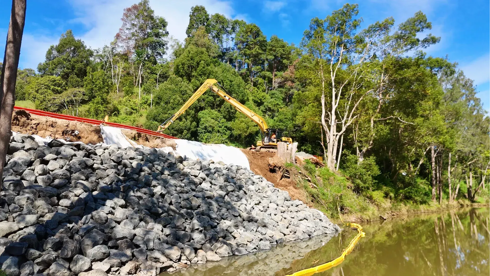 Rock riprap slope beside a calm river, with a yellow excavator arm and dense green trees under a blue sky