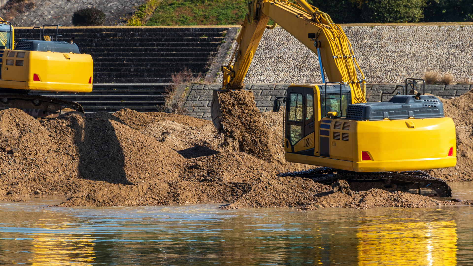 Two yellow excavators digging dirt by a riverbank