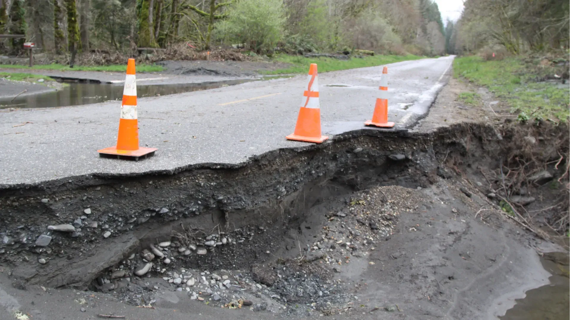 Collapsed rural road with orange traffic cones marking a large washout and eroded asphalt edge
