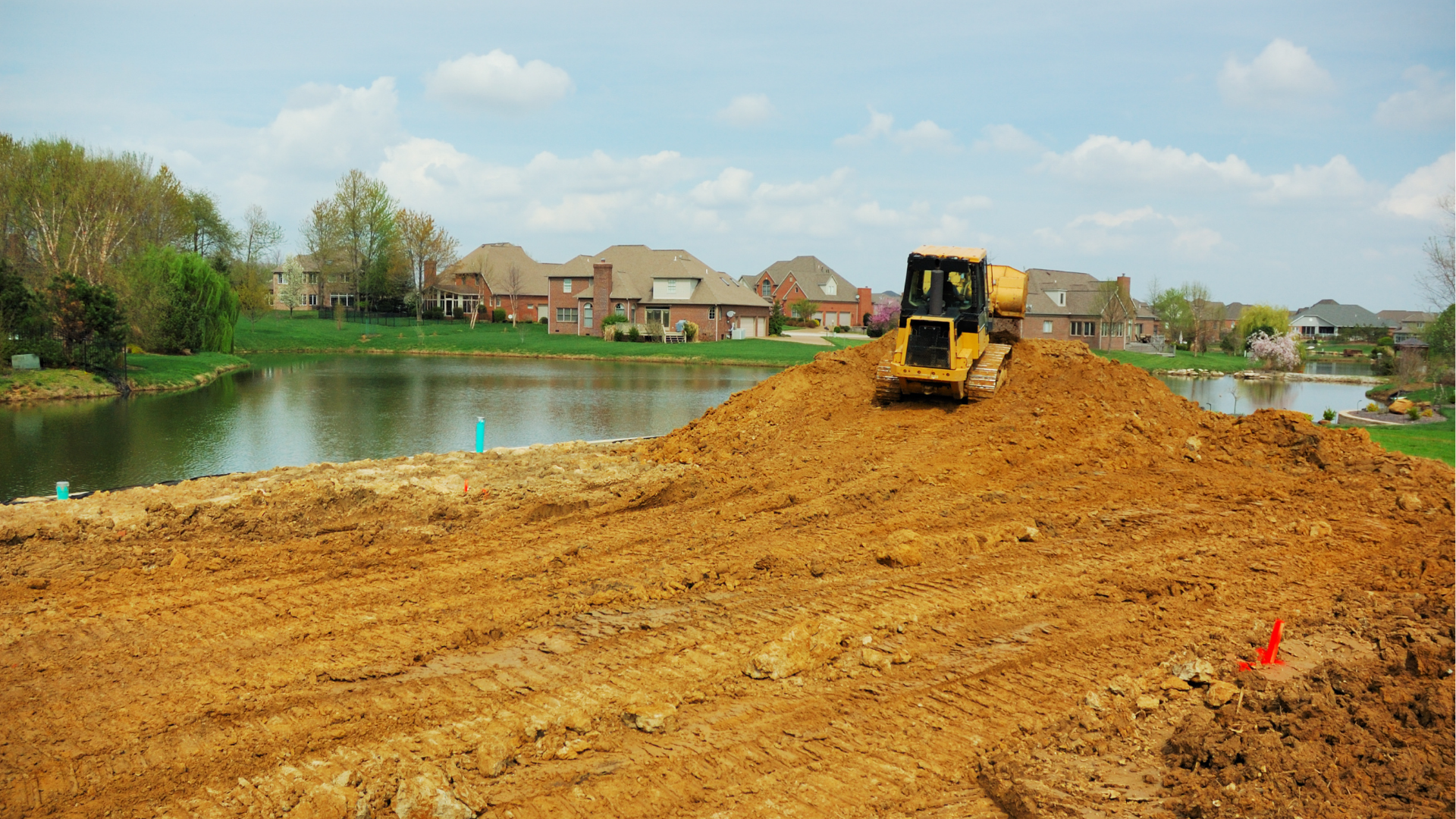 Bulldozer on a dirt embankment beside a lake, with houses and trees in the background