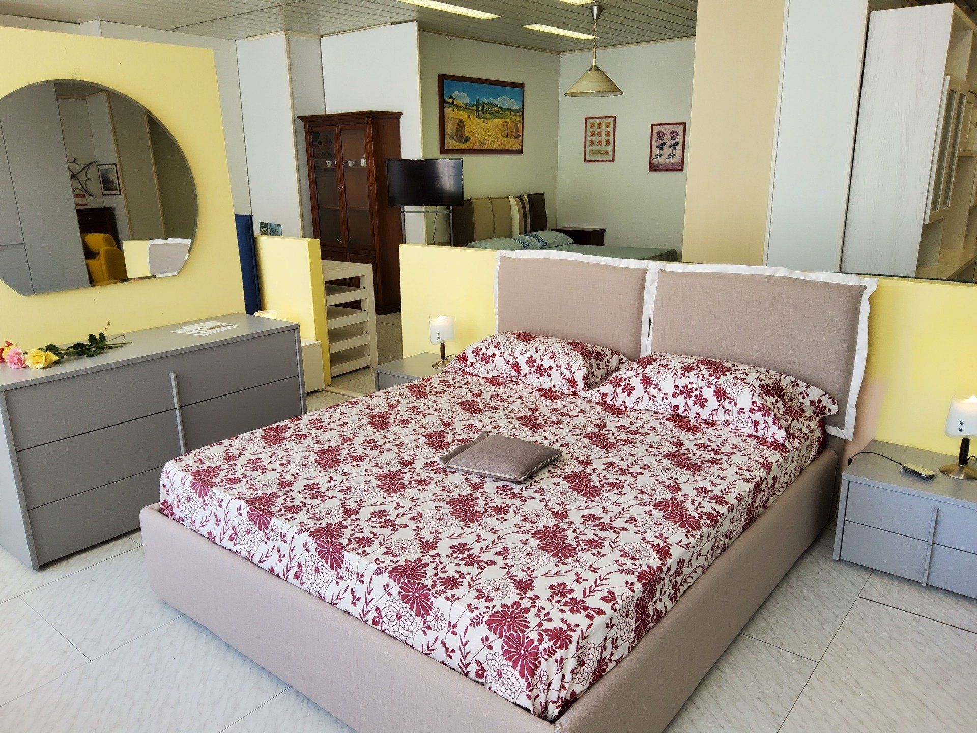 Esposizione di mobili per la camera da letto: letto con biancheria floreale, comodini, cassettiera con specchio, pareti gialle.
