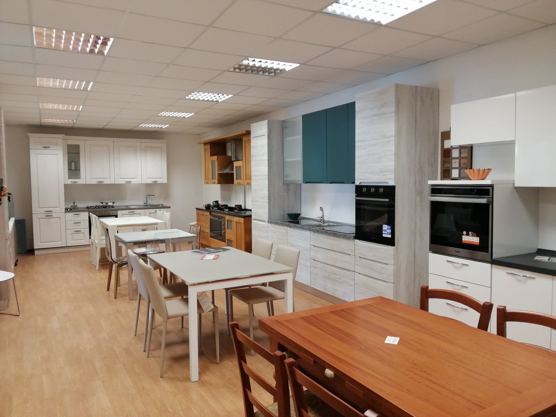 Sala espositiva della cucina con vari mobili e tavoli da pranzo. Illuminazione intensa, legno e toni neutri.