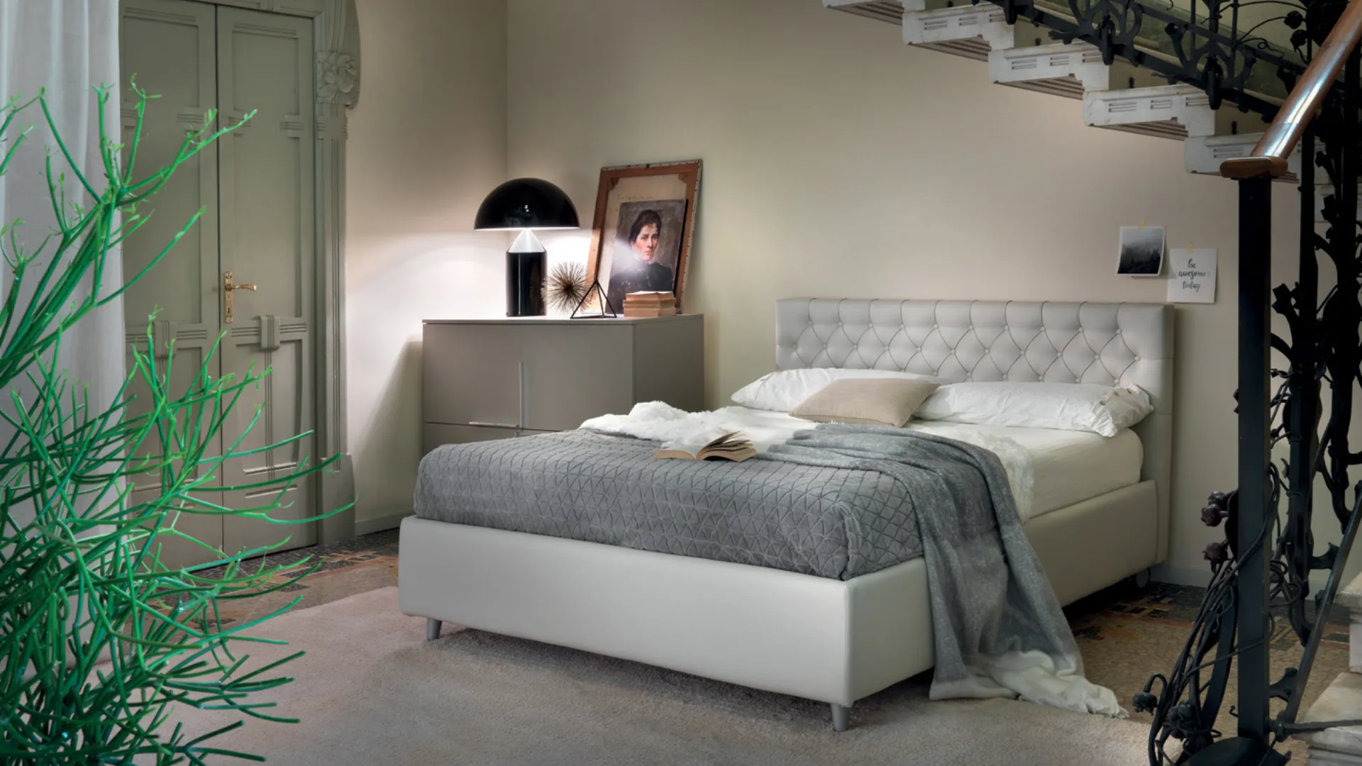Camera da letto con letto, comodino, lampada e scale. Colori grigio e bianco, luci soffuse.