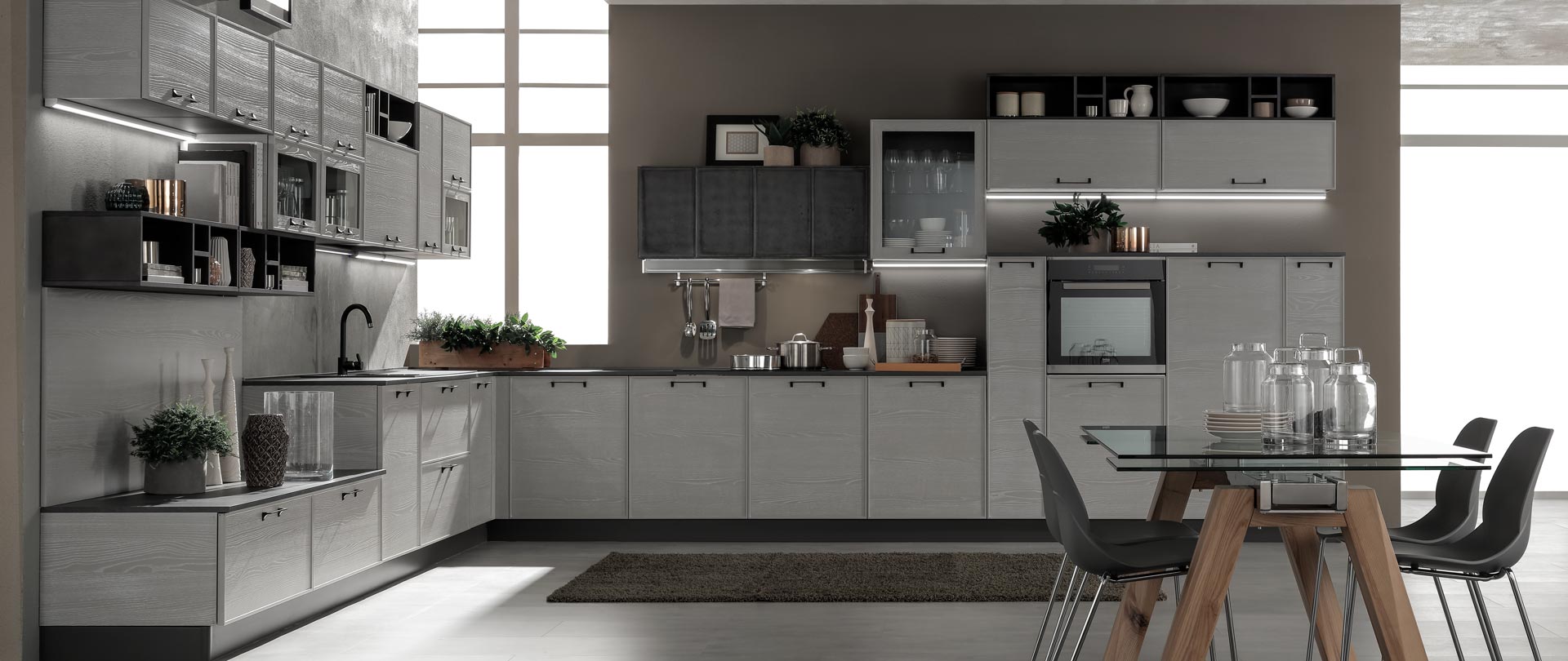 Cucina moderna con mobili grigi, forno incorporato e tavolo da pranzo con piano in vetro e sedie nere.