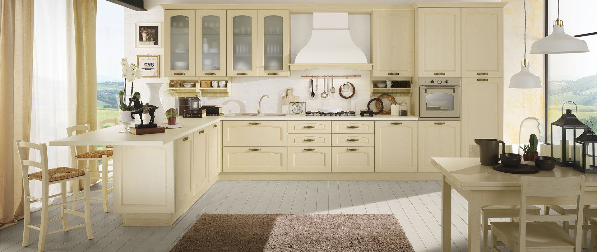 Cucina color crema con isola e tavolo da pranzo. La luce del sole entra da una grande finestra con vista sul paesaggio.